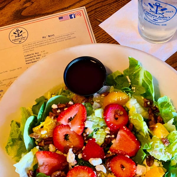 A fresh salad with strawberries, oranges, pecans, and feta cheese, with a Captain John's menu and water glass.