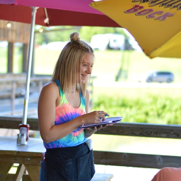 A smiling Captain John's server in a tie-dye shirt takes an order on an outdoor patio.