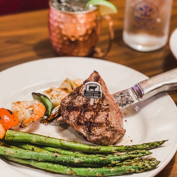 Captain John's surf and turf with steak, shrimp skewer, and asparagus on a white plate.