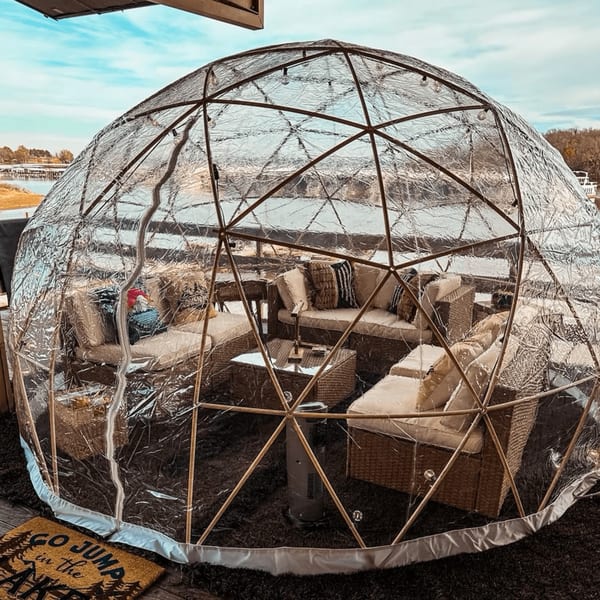 A clear geodesic dome with wicker furniture and cushions offers outdoor dining by the water.