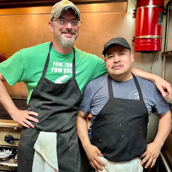 Two male chefs in aprons and hats pose in a Captain John's seafood restaurant kitchen.