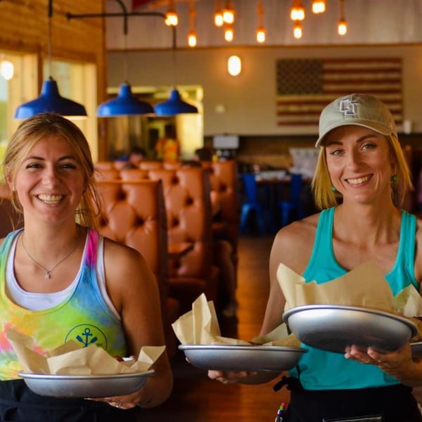 Two smiling Captain John's waitresses hold trays of food in a rustic restaurant with an American flag.
