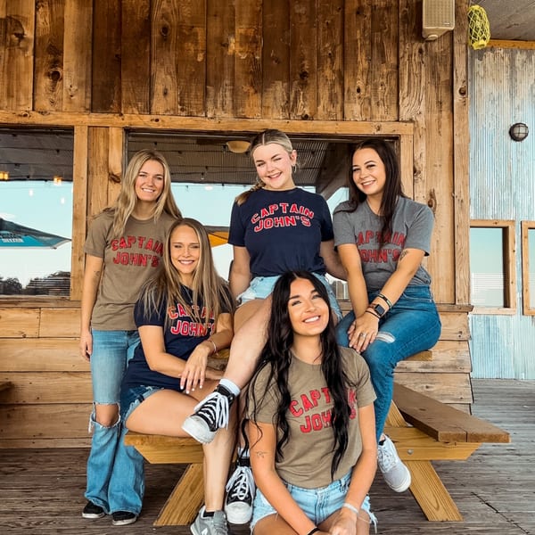 Five smiling young women wearing Captain John's t-shirts pose on a wooden bench.