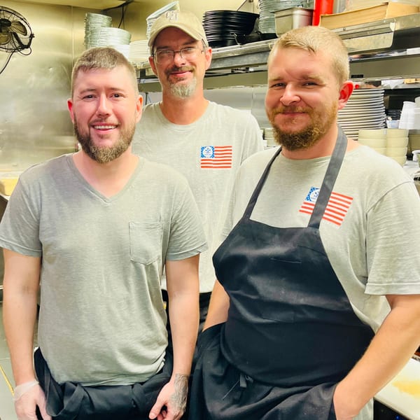 Three smiling male chefs in gray shirts and aprons stand in a Captain John's kitchen.