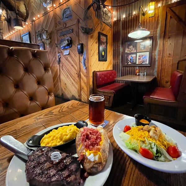 A Captain John's steak dinner with mac and cheese, baked potato, salad, and beer on a wooden table.