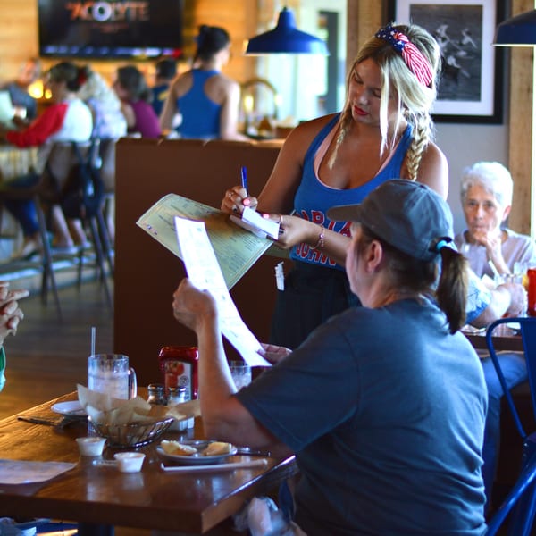 A Captain John's server in a blue tank top takes an order from a customer at a wooden table.