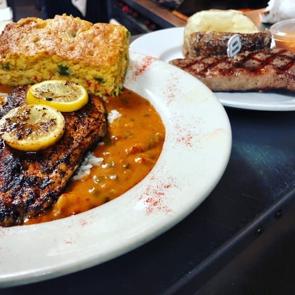 Blackened fish with lemon slices, rice, and sauce, served with cornbread and a steak.