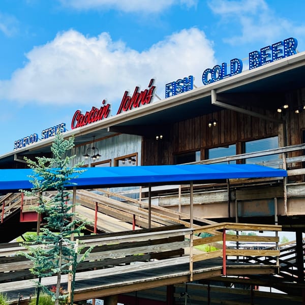 Captain John's seafood restaurant exterior with neon signs, blue awning, and wooden ramps under a blue sky.