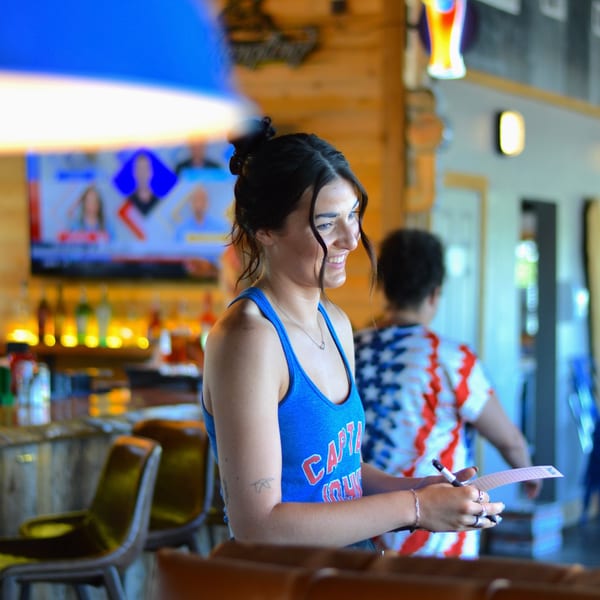 A smiling Captain John's server in a blue tank top holds a pen and paper at the bar.