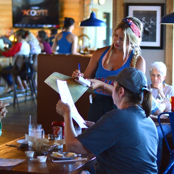 A Captain John's waitress in a blue tank top takes an order from a customer at a wooden table.