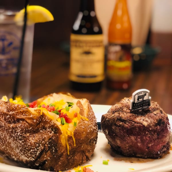 Captain John's steak and loaded baked potato, with drinks and sauces blurred in the background.