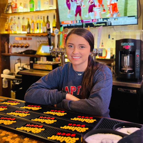 Smiling bartender in a Captain John's sweatshirt behind a bar with bottles and a TV.