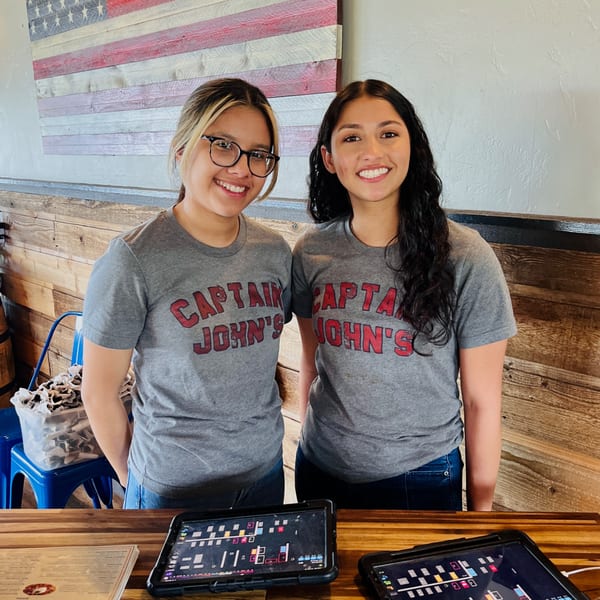Two smiling Captain John's employees in gray shirts stand behind a counter with tablets.