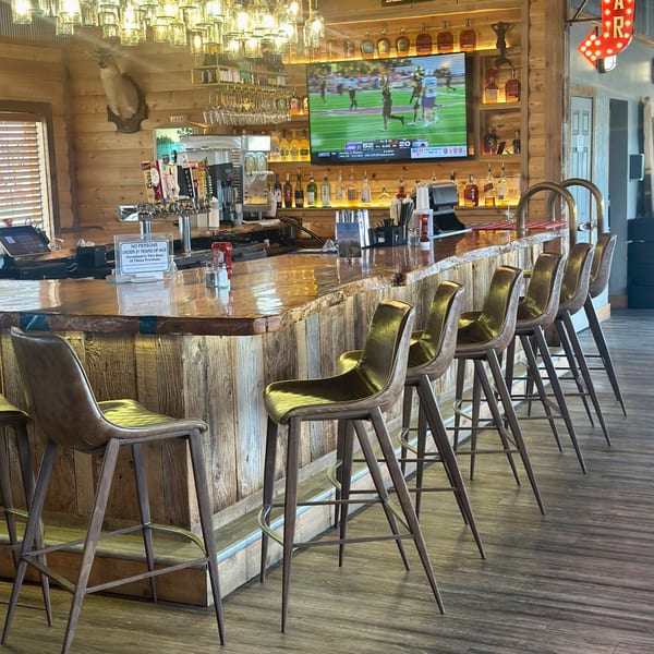 Rustic wooden bar with several stools, a large TV showing sports, and bottles of liquor.