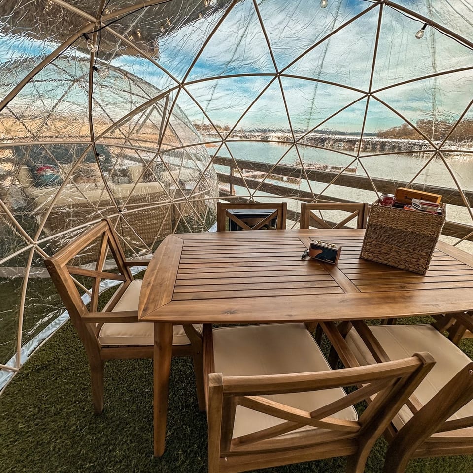 Outdoor dining igloo with a wooden table and chairs, overlooking a body of water.