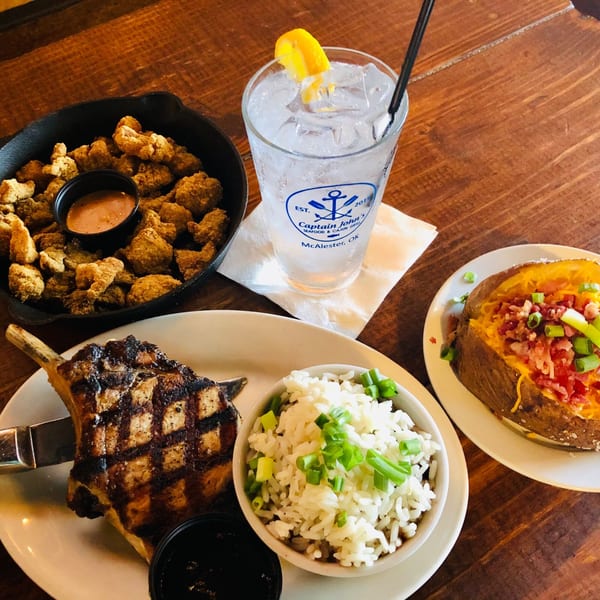 Captain John's grilled pork chop, fried oysters, rice, and loaded baked potato on a wooden table.