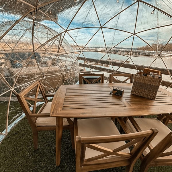 Dining table and chairs inside a clear geodesic dome, overlooking a body of water.