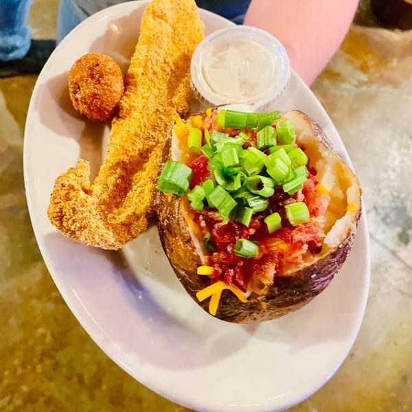 Fried fish, a hushpuppy, and a loaded baked potato with green onions and bacon on a white plate.