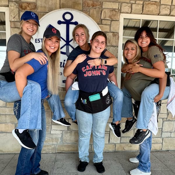 Six smiling Captain John's staff members in uniform, three giving piggyback rides to three others.