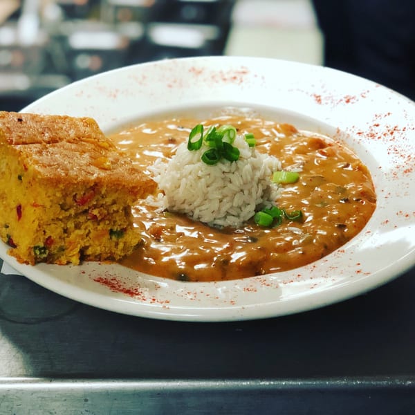 A white bowl of Captain John's seafood gumbo with rice, green onions, and a side of cornbread.